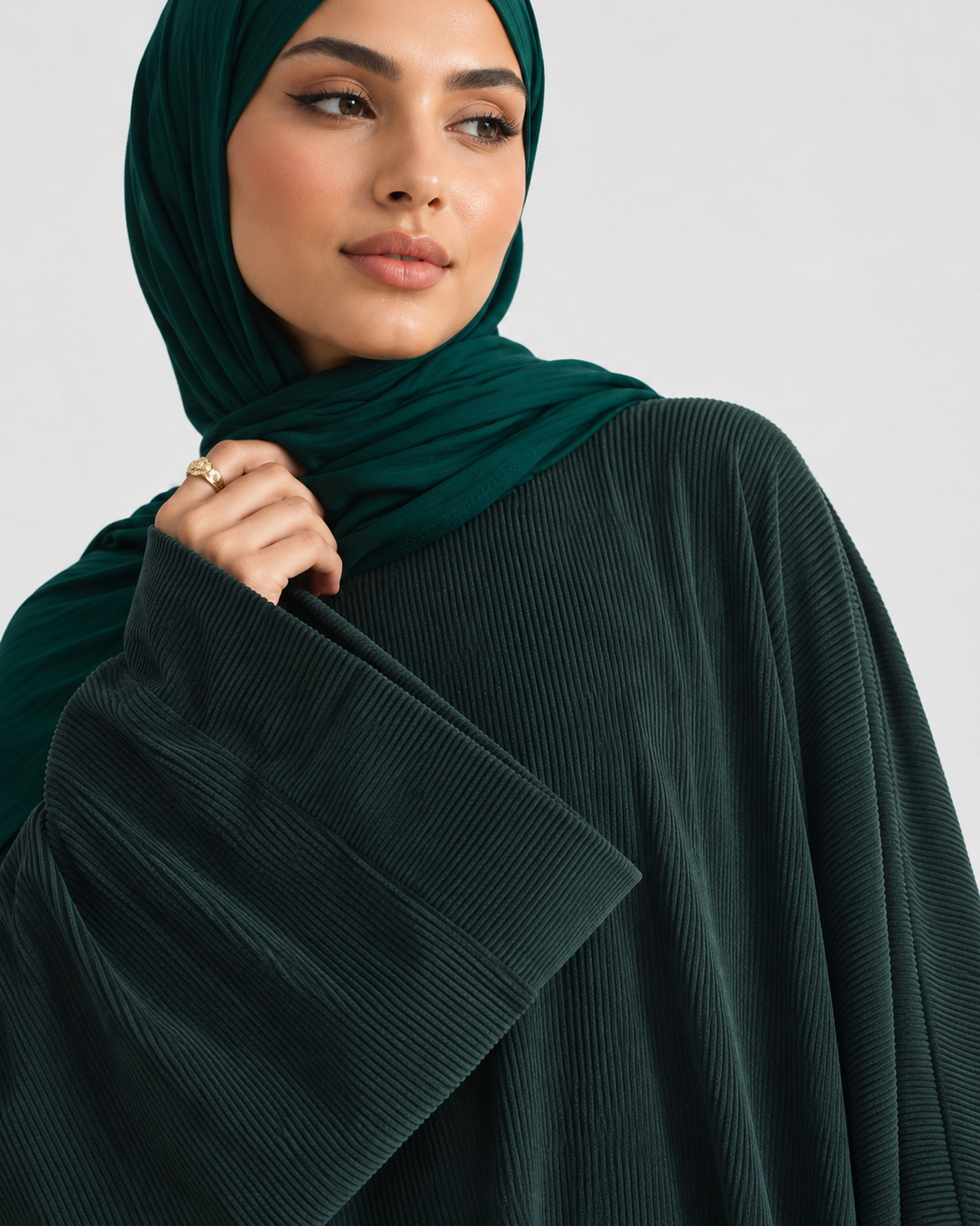 A close-up of a woman wearing a dark forest green hijab and a matching dark green pleated abaya with wide draped sleeves, set against a plain light gray background.