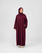 A woman modeling a long burgundy abaya with a matching hijab, standing against a light gray studio background.