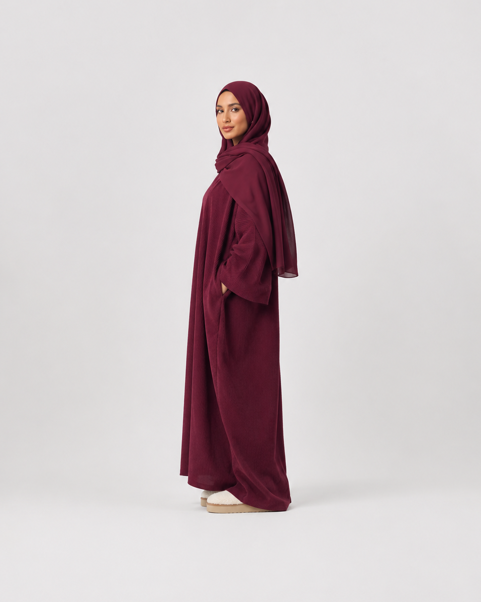 A woman wearing a deep burgundy abaya and matching hijab stands in profile against a plain light gray studio background.
