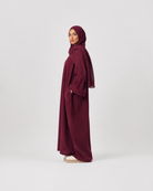 A woman wearing a deep burgundy abaya and matching hijab stands in profile against a plain light gray studio background.