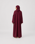 A woman wearing a long maroon abaya and matching hijab, shown from the back against a plain light gray studio background.