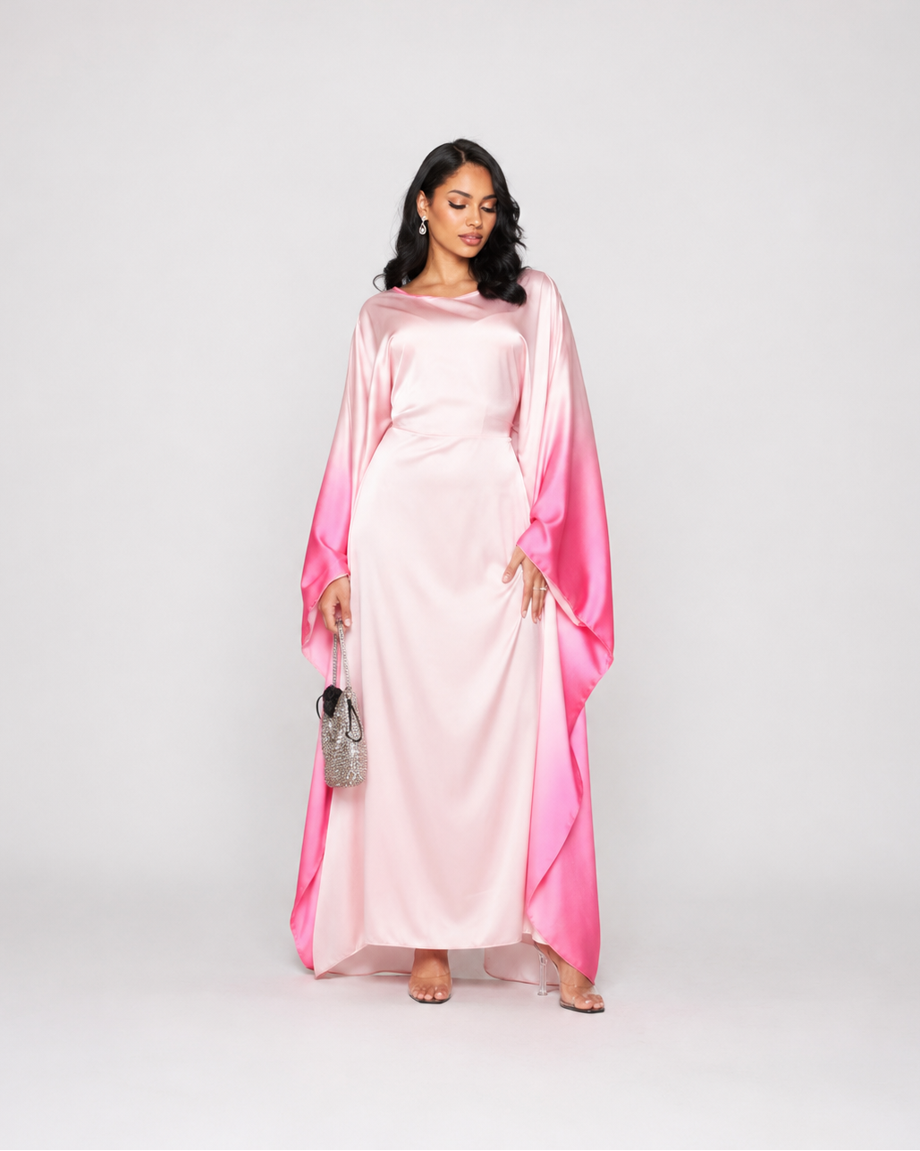 A woman modeling a long, flowing satin dress with a light pink body and vibrant pink draped sleeves, standing against a plain light gray studio background.