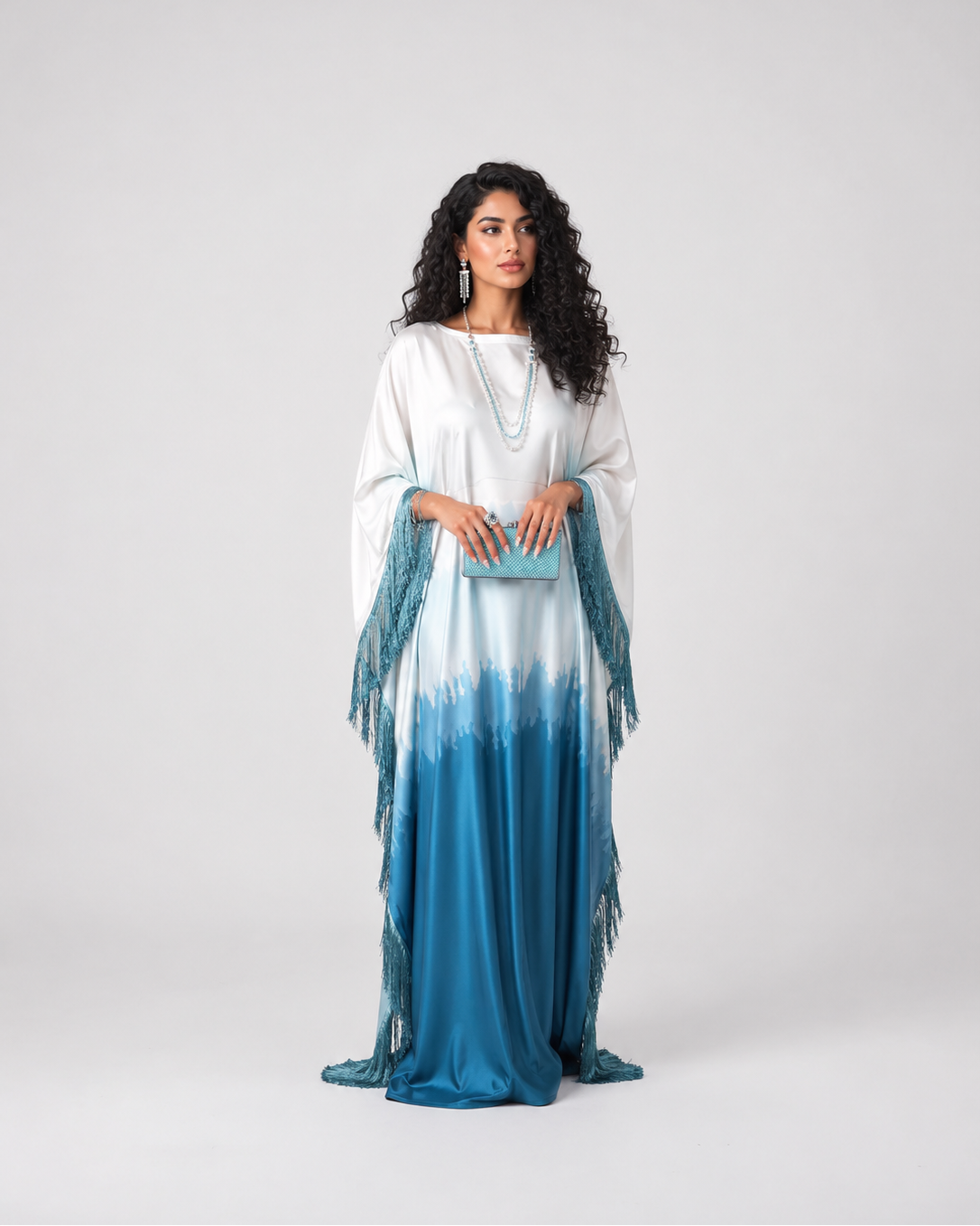 A woman modeling a long white and blue tie-dye maxi dress with fringe details on the sleeves, standing against a light gray studio background.