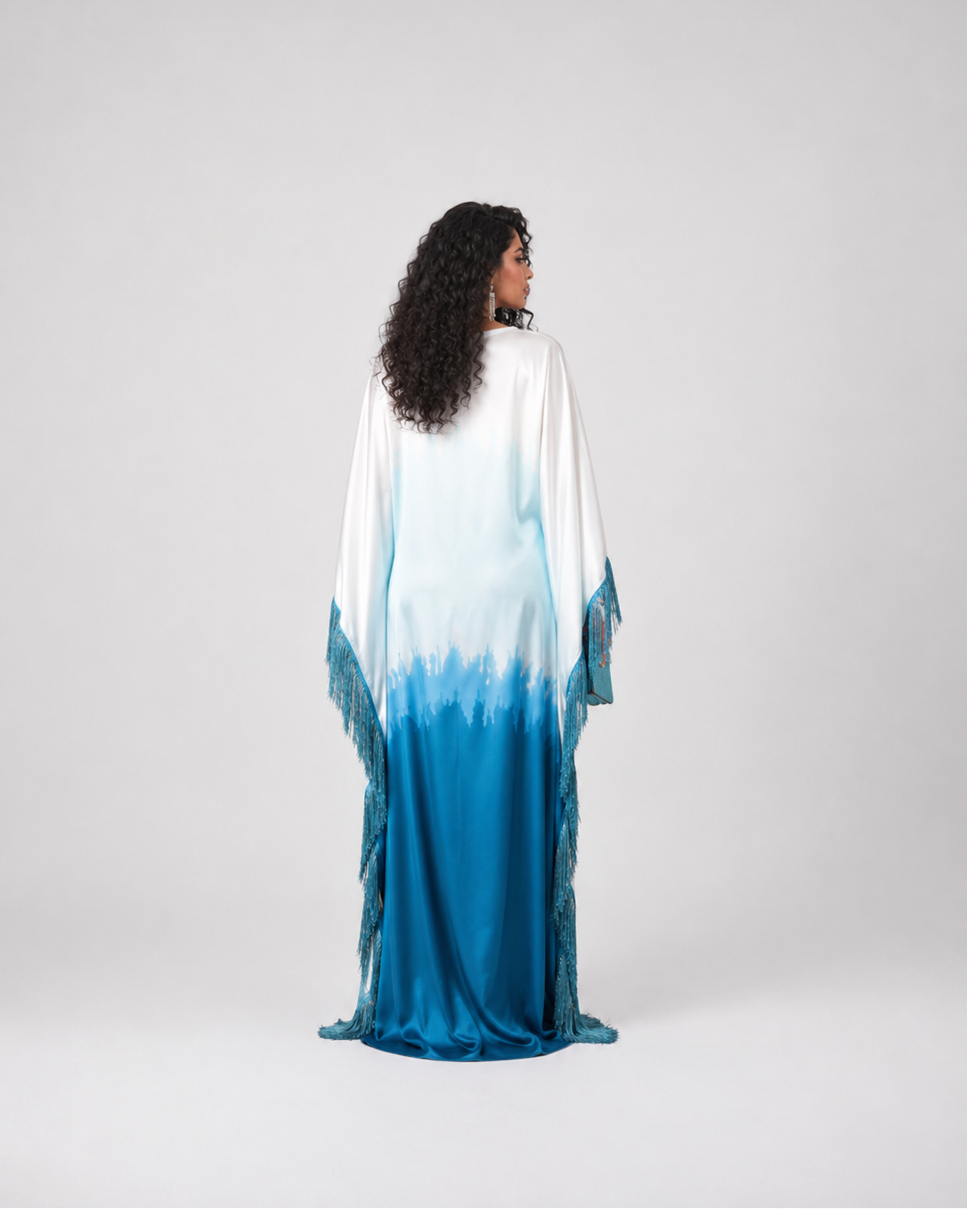 A woman stands with her back to the camera wearing a long flowing kaftan dress with a white-to-blue ombré gradient and fringe details, photographed against a light gray studio background.