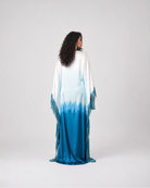 A woman stands with her back to the camera wearing a long flowing kaftan dress with a white-to-blue ombré gradient and fringe details, photographed against a light gray studio background.