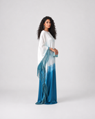 A woman modeling a long white and blue tie-dye maxi dress with fringe details, standing against a light gray studio background.