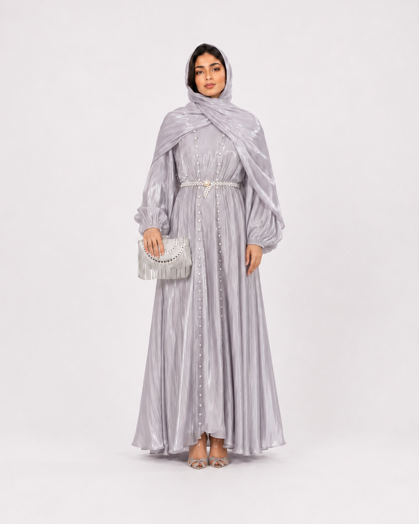 A woman modeling a long, flowing light gray maxi dress with a matching draped shawl and holding a small metallic clutch, photographed against a seamless light gray studio background.