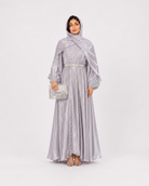 A woman modeling a long, flowing light gray maxi dress with a matching draped shawl and holding a small metallic clutch, photographed against a seamless light gray studio background.