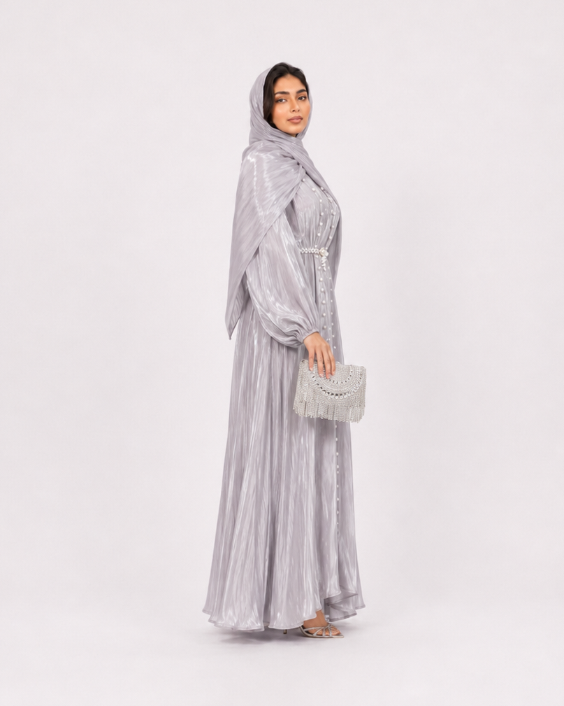 A woman modeling a light silver-gray maxi abaya with a matching headscarf and holding a small matching clutch bag, standing against a plain light gray studio background.