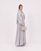 A woman modeling a light silver-gray maxi abaya with a matching headscarf and holding a small matching clutch bag, standing against a plain light gray studio background.