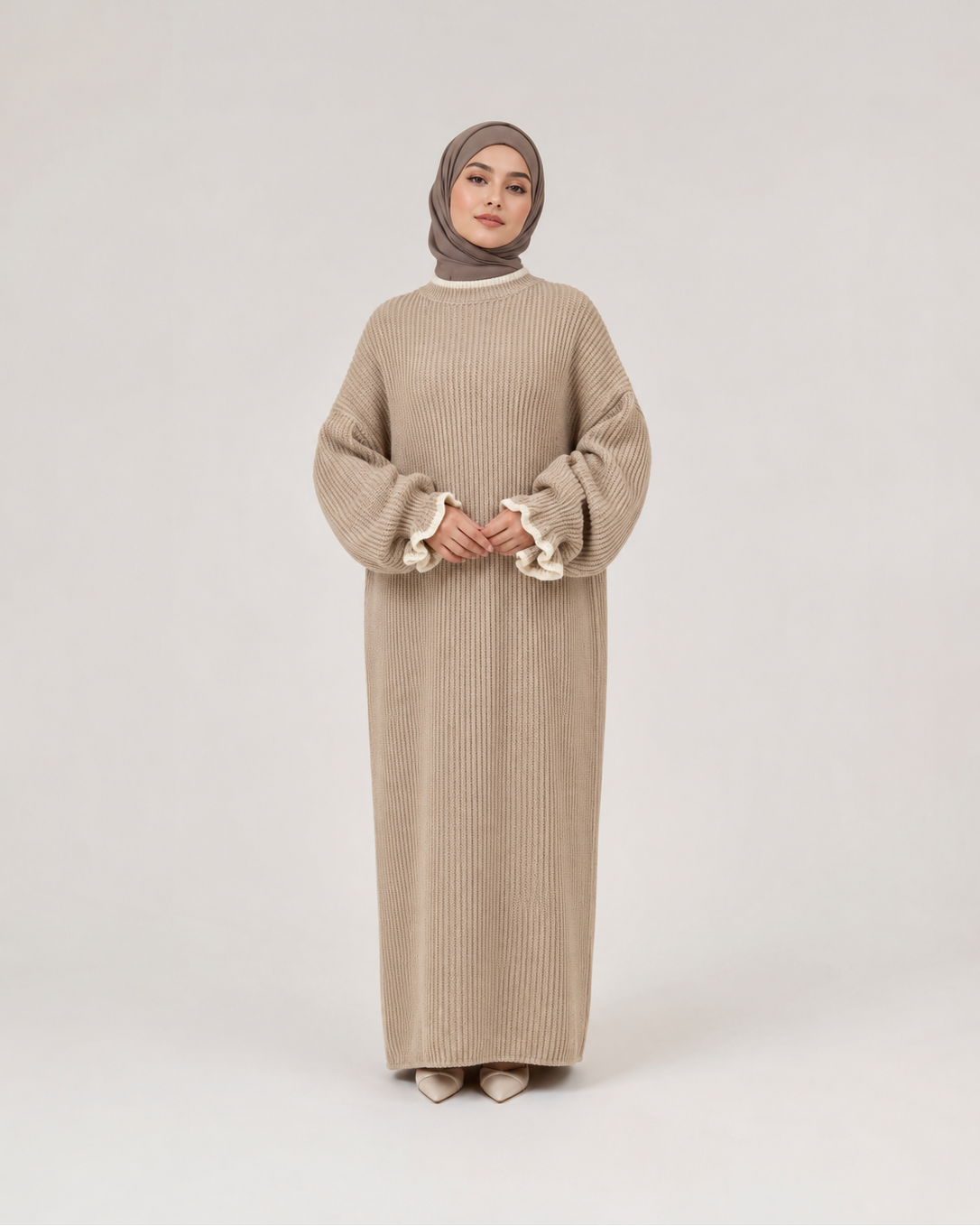A woman modeling a long beige ribbed knit abaya dress with wide sleeves and ruffled cuffs, paired with a taupe hijab, standing against a light gray studio background.