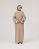 A woman modeling a long beige ribbed knit abaya dress with wide sleeves and ruffled cuffs, paired with a taupe hijab, standing against a light gray studio background.