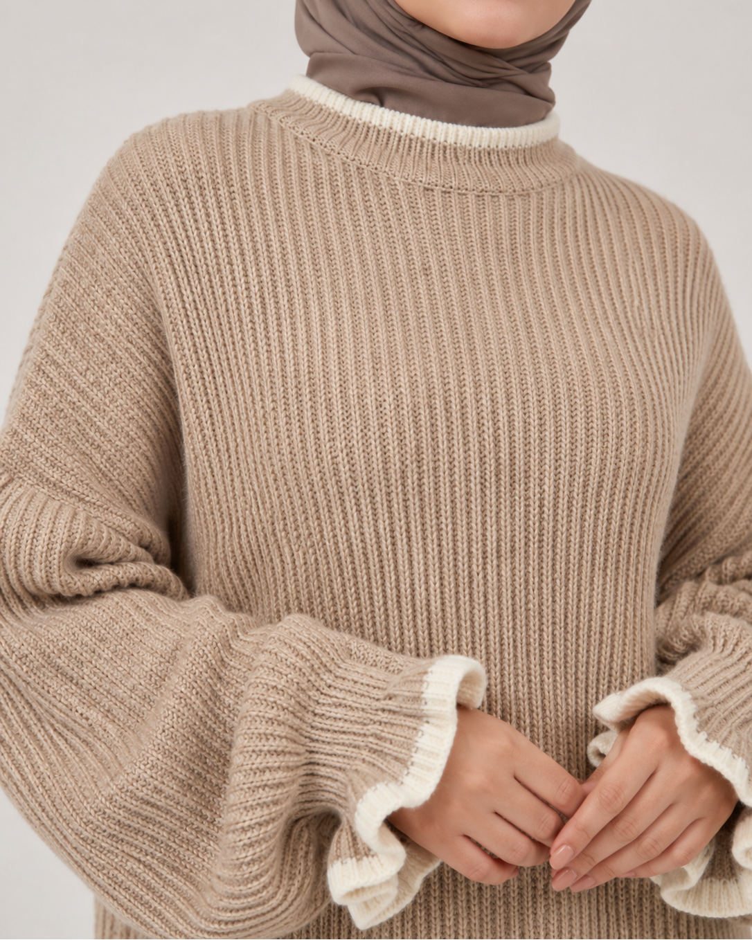 A close-up of a woman wearing a beige ribbed knit sweater with white ruffled trim at the collar and cuffs, paired with a taupe hijab, against a light gray studio background.