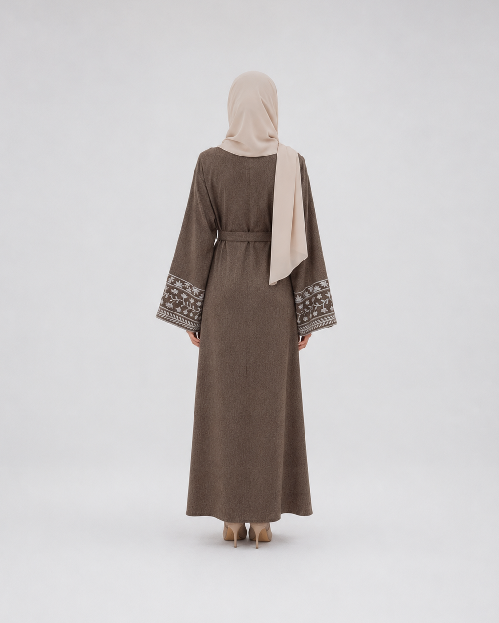 A woman viewed from behind wearing a long taupe-brown abaya with patterned embroidery on the wide sleeves and a light beige hijab, standing against a plain light gray studio background.