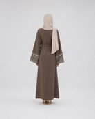 A woman viewed from behind wearing a long taupe-brown abaya with patterned embroidery on the wide sleeves and a light beige hijab, standing against a plain light gray studio background.