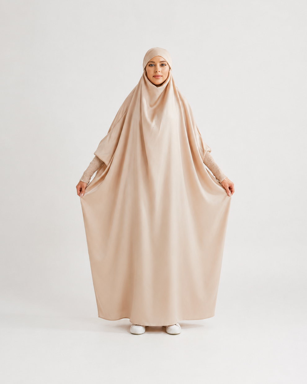 A woman wearing a loose, flowing beige hijab and long-sleeve abaya outfit, holding the fabric outwards against a plain light gray studio background.