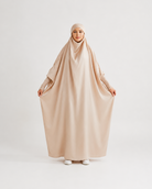 A woman wearing a loose, flowing beige hijab and long-sleeve abaya outfit, holding the fabric outwards against a plain light gray studio background.