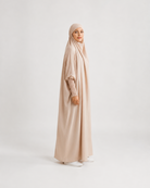 A woman wearing a long, flowing abaya in a light beige color with a matching hijab, standing in profile against a plain light gray studio background.