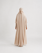 A person viewed from the back wearing a flowing light beige or blush-beige full-length garment with a matching head covering, standing against a plain light gray studio background.