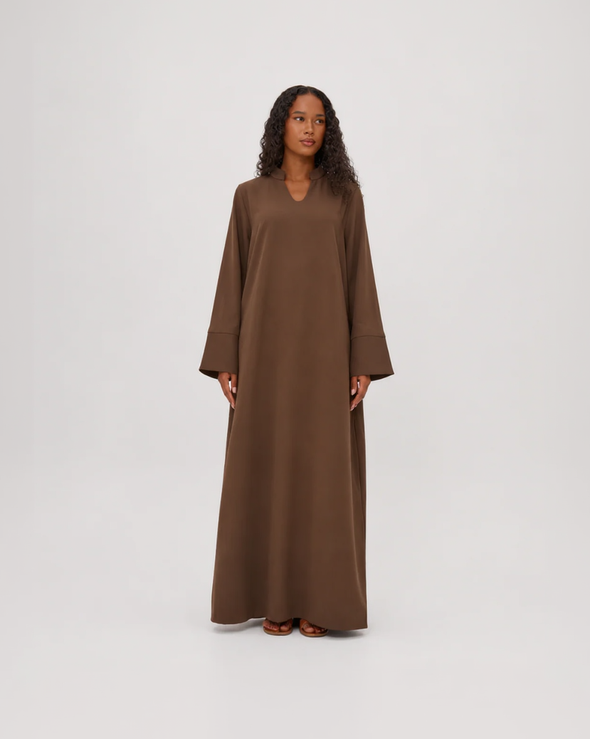 A woman modeling a long, loose-fitting brown maxi dress with wide sleeves and a V-neckline, standing against a plain light gray studio background.