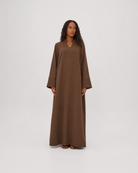 A woman modeling a long, loose-fitting brown maxi dress with wide sleeves and a V-neckline, standing against a plain light gray studio background.