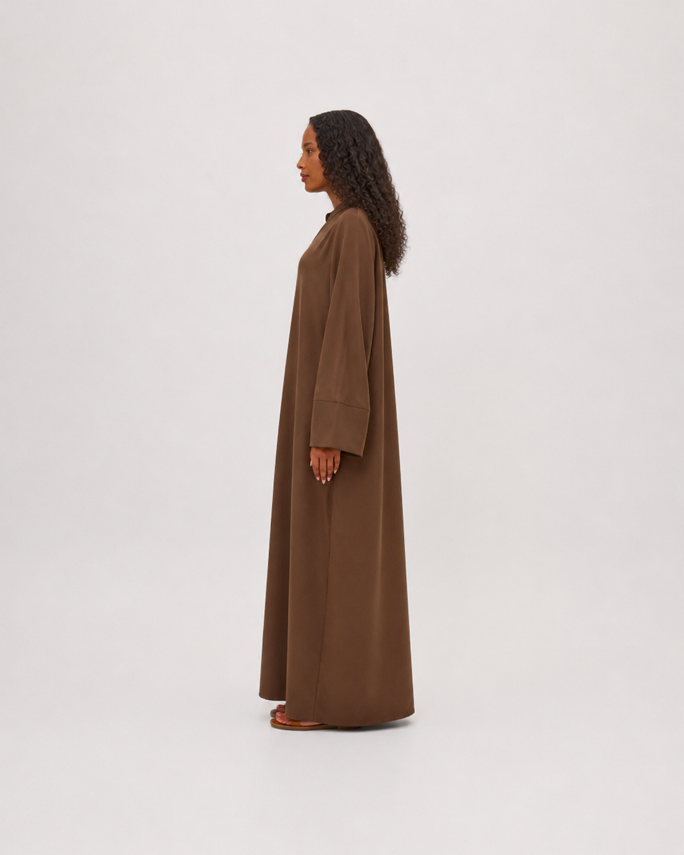 A woman stands in profile wearing a long, loose-fitting brown abaya or maxi dress against a plain light gray studio background.