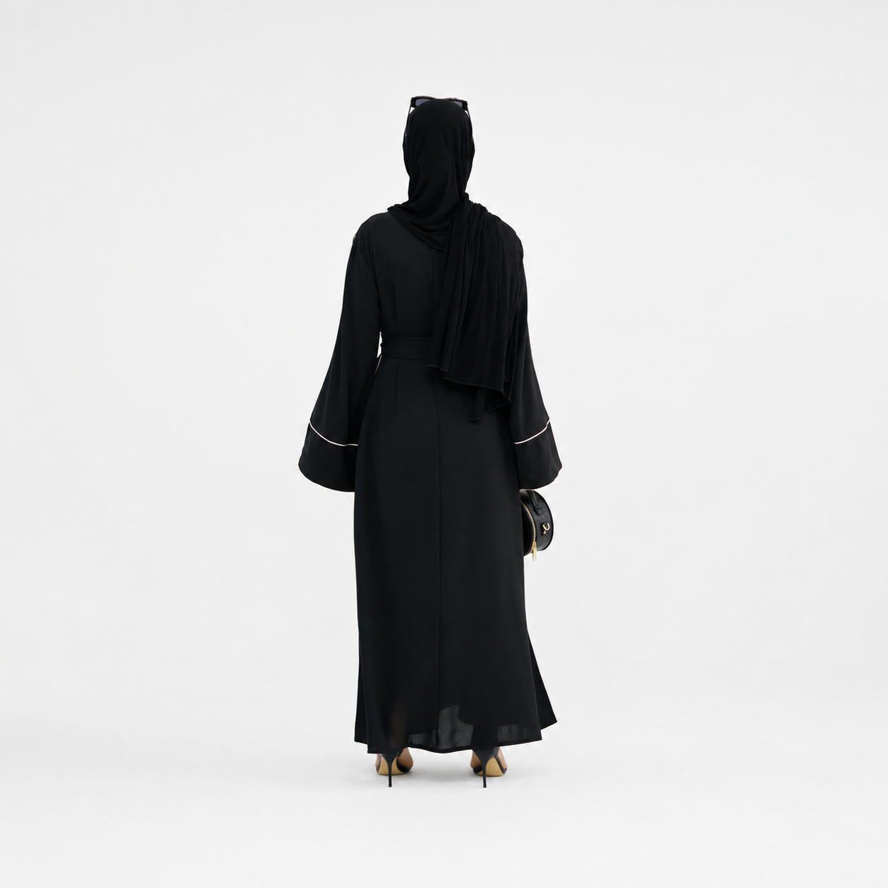 A woman viewed from behind wearing a long black abaya with wide sleeves and thin white piping details at the cuffs, along with a black hijab and black heels, against a plain white studio background.
