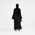 A woman viewed from behind wearing a long black abaya with wide sleeves and thin white piping details at the cuffs, along with a black hijab and black heels, against a plain white studio background.