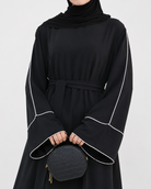 A person wearing a black long-sleeve abaya dress with white contrast piping and a matching waist tie, paired with a black headscarf and holding a small black quilted handbag against a light gray studio background.