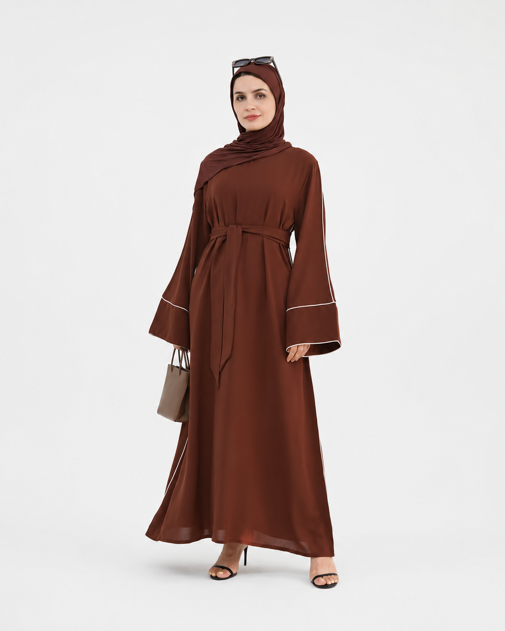 A woman modeling a long brown abaya dress with a matching headscarf, featuring wide bell sleeves with white piping details, a self-tie waist belt, and sunglasses on her head, standing against a light gray studio background.