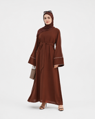 A woman modeling a long brown abaya dress with a matching headscarf, featuring wide bell sleeves with white piping details, a self-tie waist belt, and sunglasses on her head, standing against a light gray studio background.
