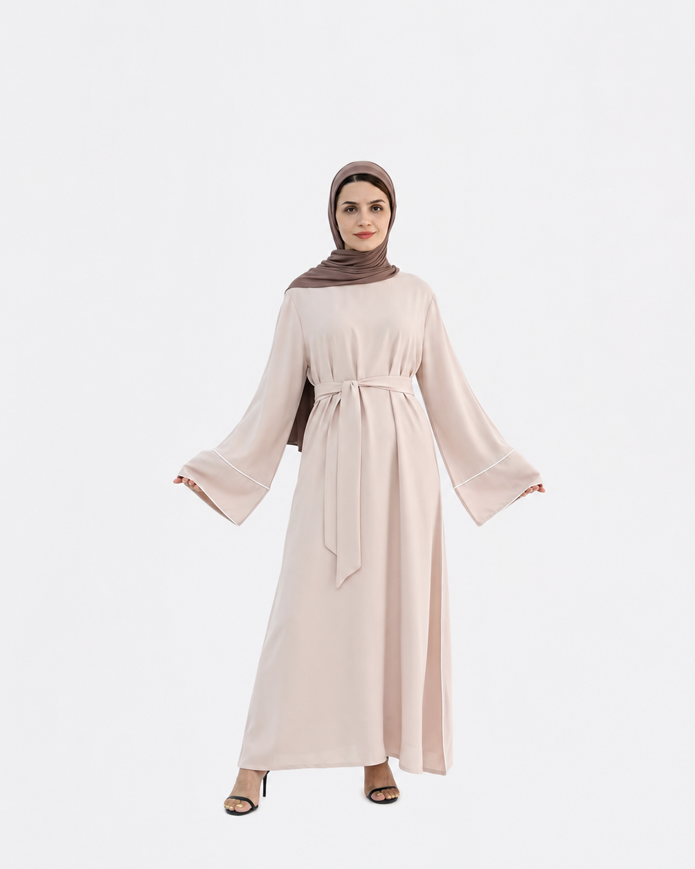 A woman modeling a long, light blush pink abaya dress with wide bell sleeves and a matching waist tie, paired with a brown hijab and black heeled sandals, set against a plain light gray background.