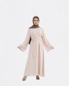 A woman modeling a long, light blush pink abaya dress with wide bell sleeves and a matching waist tie, paired with a brown hijab and black heeled sandals, set against a plain light gray background.