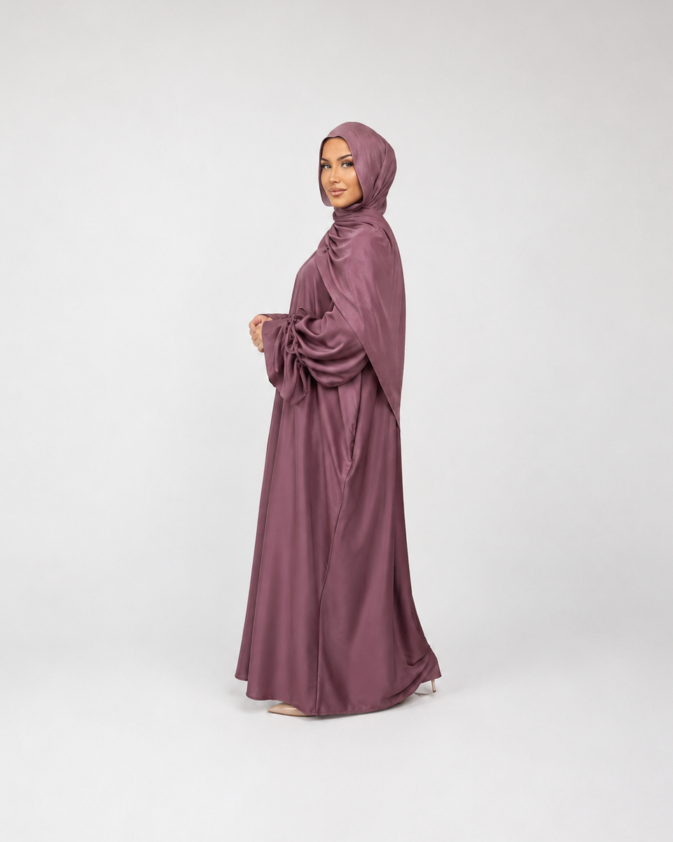 A woman wearing a flowing mauve-colored abaya and matching hijab, standing in profile against a light gray studio background.