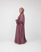 A woman wearing a flowing mauve-colored abaya and matching hijab, standing in profile against a light gray studio background.