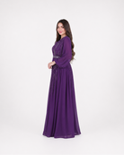 A woman modeling a long, floor-length purple gown with long sleeves and a fitted waist, standing against a plain light gray studio background.