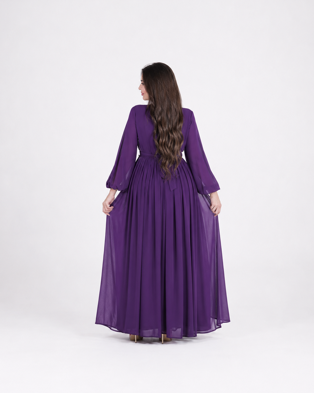 A woman viewed from behind wearing a long purple maxi dress with three-quarter sleeves and a gathered waist, standing against a plain light gray studio background.