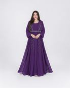 A woman modeling a floor-length purple gown with long sleeves, a square neckline, and silver beaded embellishments, standing against a light gray studio background.