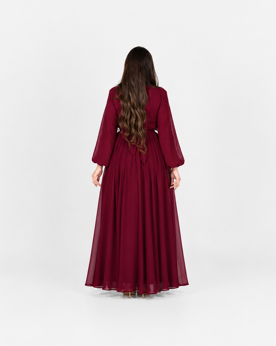 A woman viewed from behind wearing a long, flowing burgundy dress with three-quarter sleeves, standing against a plain light gray studio background.