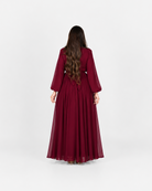 A woman viewed from behind wearing a long, flowing burgundy dress with three-quarter sleeves, standing against a plain light gray studio background.