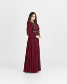 A woman modeling a long, floor-length maroon dress with long sleeves, a fitted waist, and embroidered detailing on the bodice, standing against a plain light gray studio background.