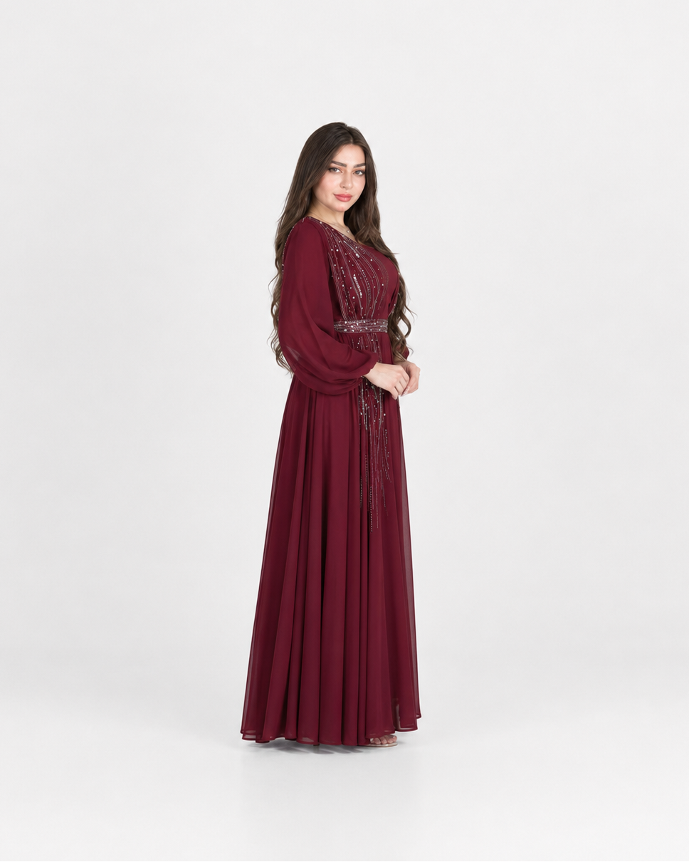 A woman modeling a long, floor-length maroon dress with long sleeves, a fitted waist, and embroidered detailing on the bodice, standing against a plain light gray studio background.
