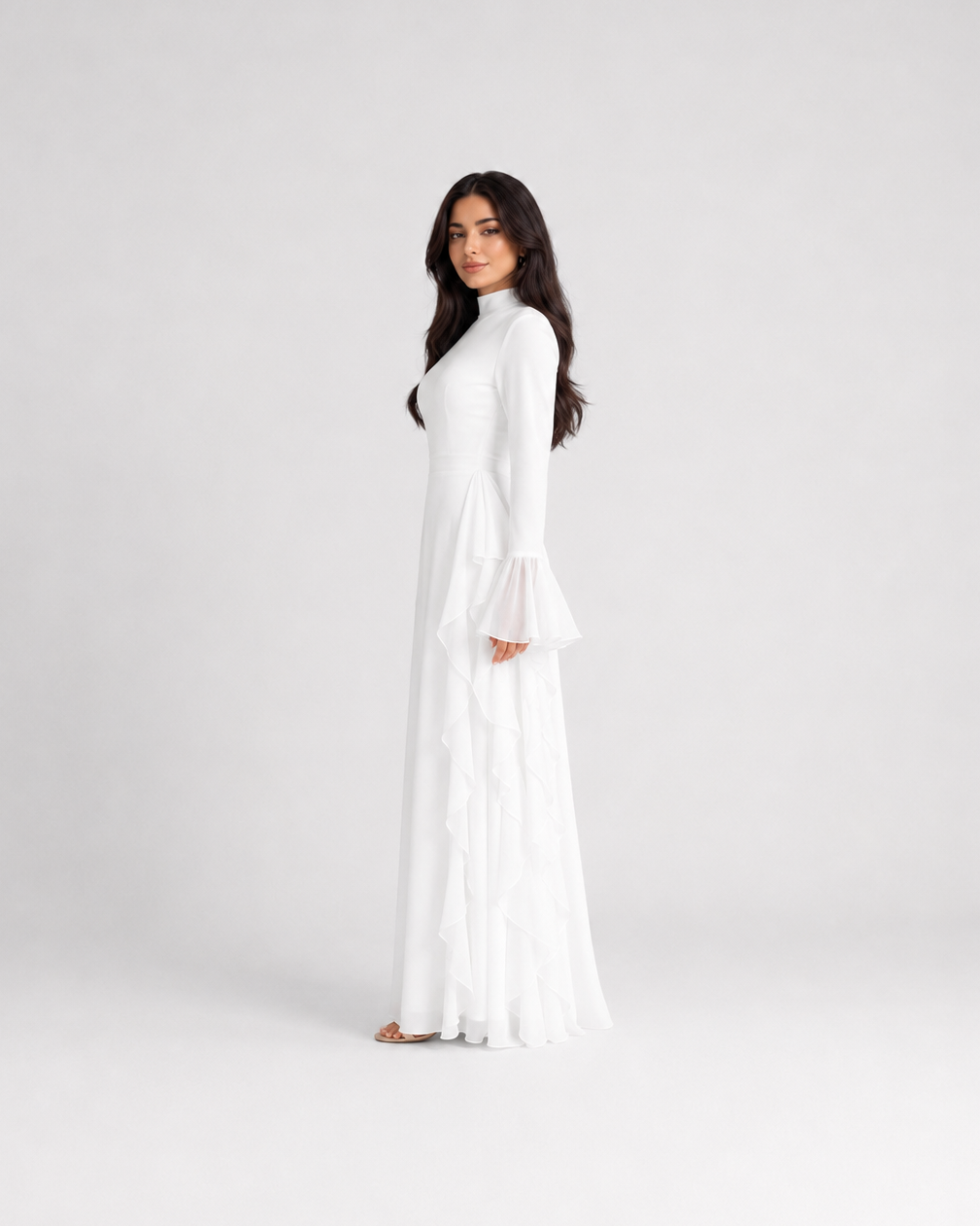 A woman modeling a long white maxi dress with long bell sleeves and a high neckline, standing against a plain light gray studio background.