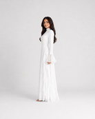 A woman modeling a long white maxi dress with long bell sleeves and a high neckline, standing against a plain light gray studio background.