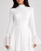 A woman wearing a white long-sleeve mock-neck dress with flared bell sleeves and a high-low layered hem, photographed against a light gray studio background.
