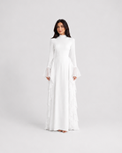 A woman modeling a long-sleeved white maxi dress with tiered ruffle details along the skirt, standing against a seamless light gray studio background.