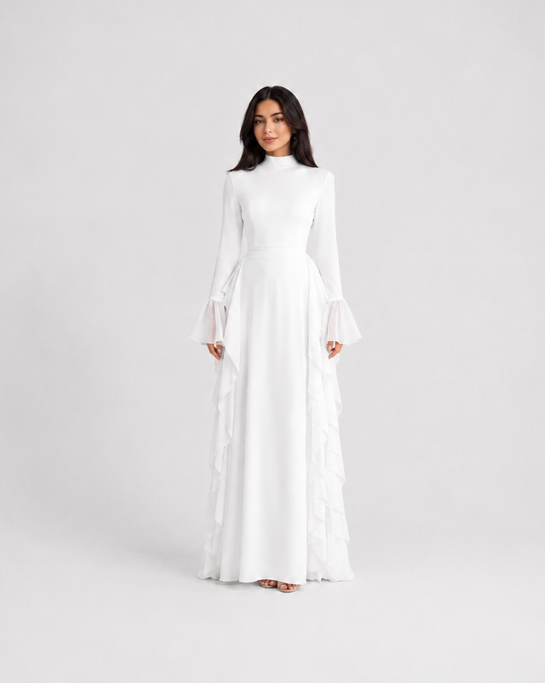 A woman modeling a long-sleeved white maxi dress with tiered ruffle details along the skirt, standing against a seamless light gray studio background.