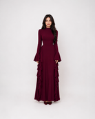 A woman modeling a long-sleeved, high-neck maxi dress in deep burgundy with dramatic ruffled layers on the skirt, set against a plain light gray studio background.