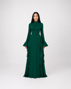 A woman modeling a dark green long-sleeve maxi dress with a high mock neck, flared bell sleeves, and a tiered ruffle skirt, set against a plain light gray studio background.