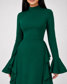 A woman wearing a dark green long-sleeve dress with a high mock neck, fitted waist, and dramatic flared ruffled bell sleeves, shown against a light gray studio background.